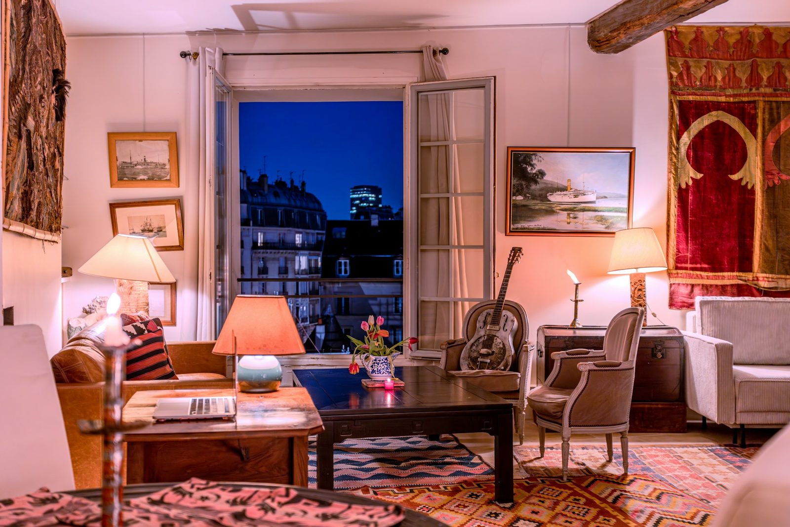 Séjour d'un appartement ancien le soir, fauteuils de style et tapis, porte-fenêtre ouverte sur les toits d'immeubles et une tour éclairée