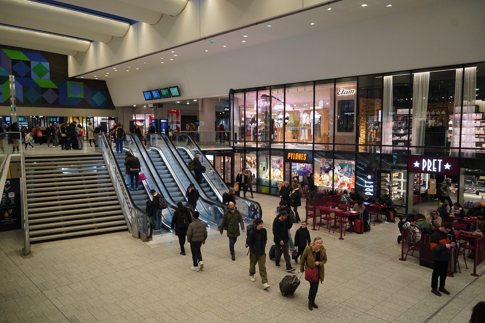 Voyageurs avec valises devant les escaliers et escalators de la gare Montparnasse, bordés de boutiques et d'une terrasse de café
