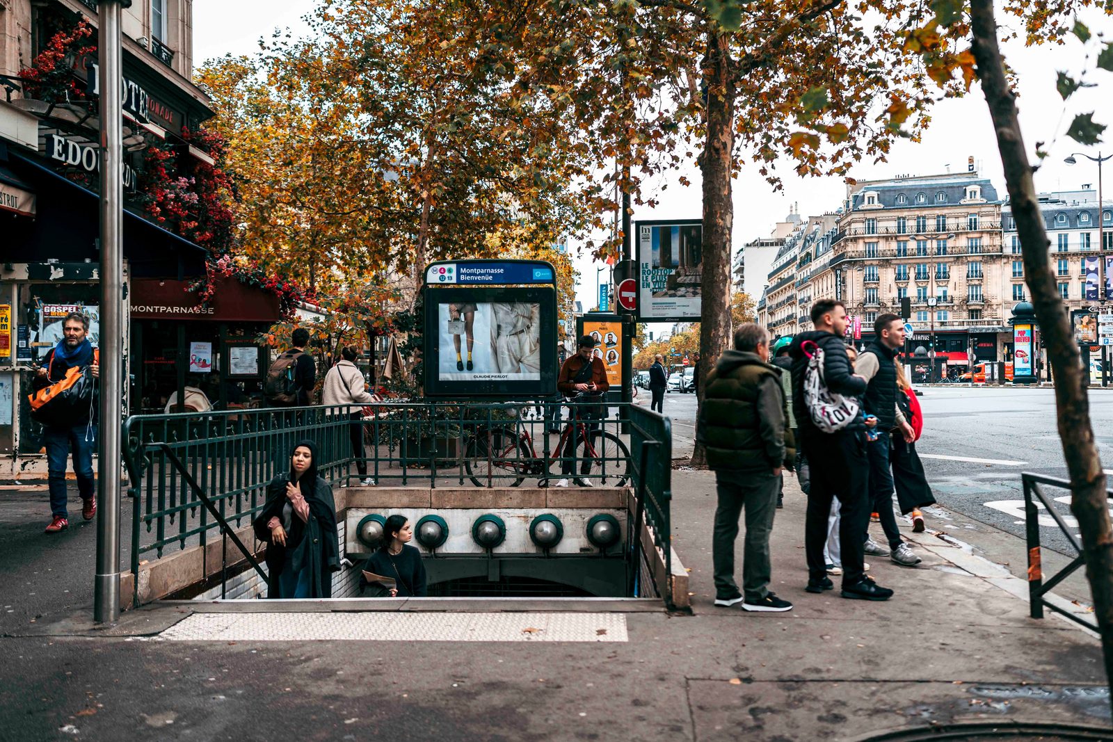 Bouche de métro surmontée de la plaque Montparnasse-Bienvenüe, sur un trottoir arboré en automne, des passants autour
