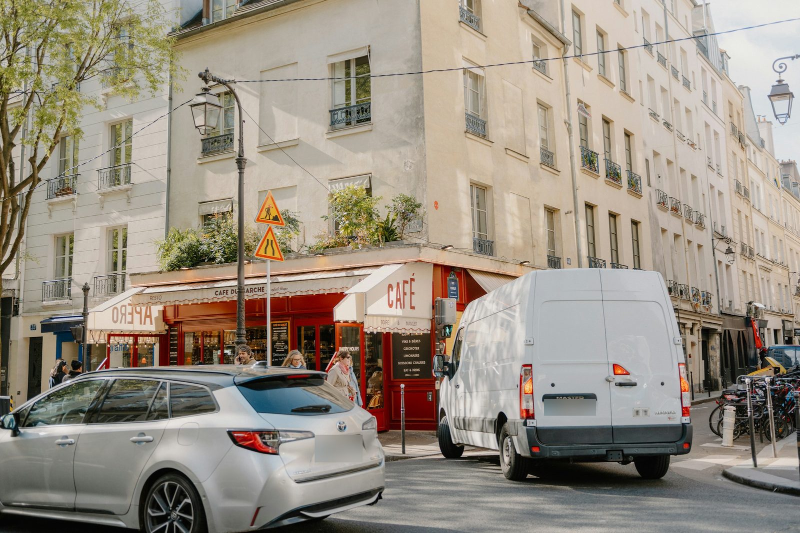 Fourgon Renault Master blanc arrêté à un carrefour parisien devant un café à devanture rouge, à côté d'une voiture