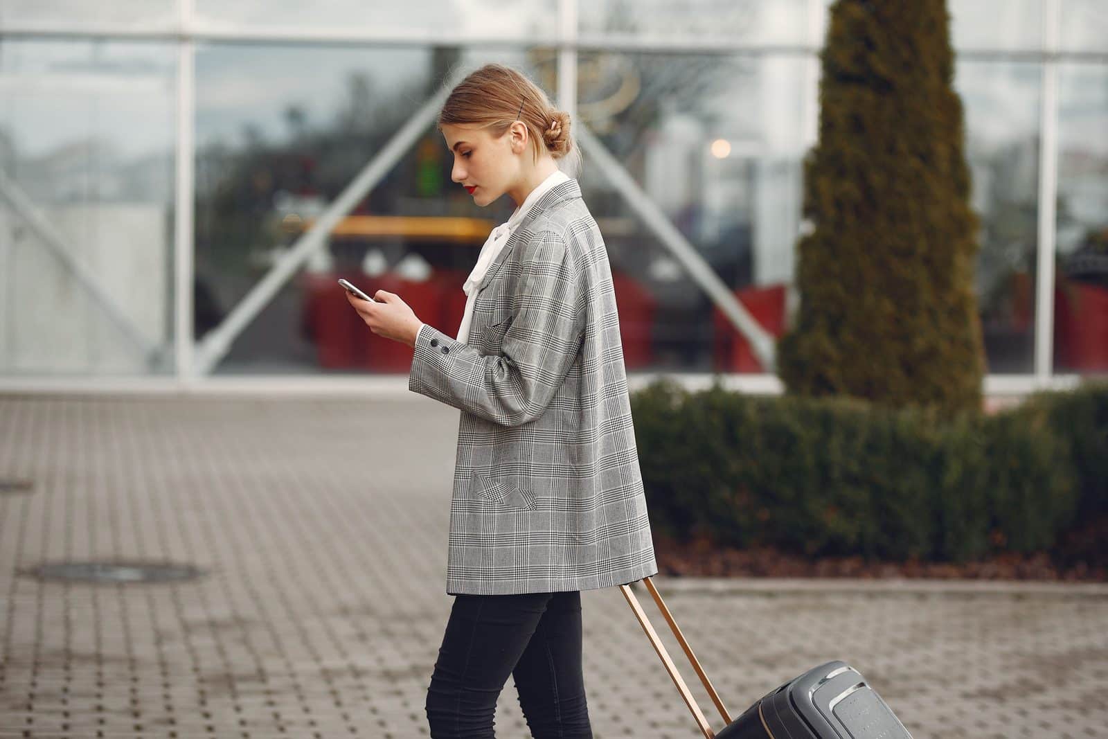 Voyageuse tirant une valise à roulettes et consultant son smartphone devant une façade vitrée